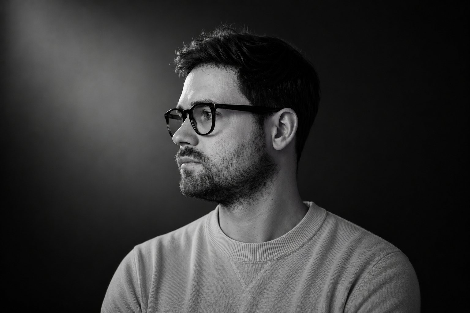 Schwarzweiss-Portrait eines Mannes mit Brille mit Blick nach links vor dunklem Hintergrund, Licht von links oben